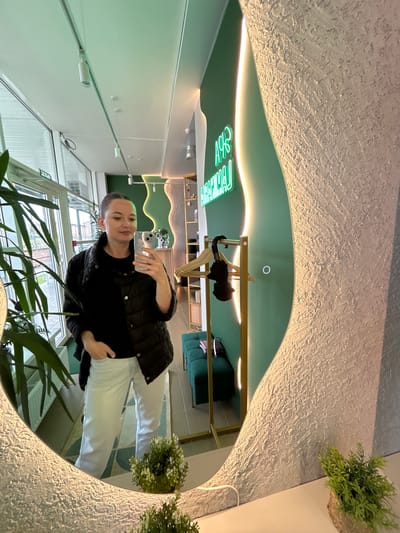 A woman with brown hair pulled back in a bun, wearing a black puffer jacket over a black t-shirt and white pants, stands in front of a large, irregularly shaped mirror in what appears to be a modern, clean establishment. The mirror reflects the woman and the interior of the space, which is painted in shades of green. A neon sign with Russian text, "АРТ-ЛАУНЖ", is visible on the green wall. The environment includes large windows, plants, a gold clothing rack with a black umbrella, and green seating. The overall impression is a well-lit, stylish interior, possibly a salon or lounge area, located in Medvedevo Urban Settlement, Russia.