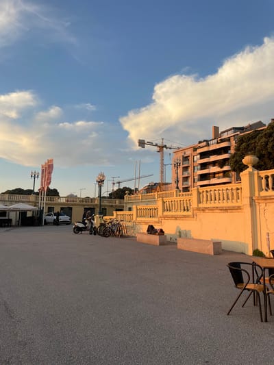 This image depicts an outdoor public area, likely a promenade, in Aldoar, Foz do Douro e Nevogilde, Portugal, under a clear blue sky with scattered, elongated white clouds, illuminated by warm, low-angle sunlight originating from the left. The ground surface is dark asphalt. Partial views of dark outdoor cafe chairs and a table are visible on the extreme right.
In the midground, a light-yellow concrete retaining wall with a decorative balustrade extends horizontally, exhibiting visible cracks in its surface. Two light-colored, rectangular concrete benches are positioned along this wall. One individual is observed lying prone on the left bench, partially covered by dark fabric and a light brown object. Another person is seated on the ground adjacent to the right bench, partially obscured. To the left of the benches, several bicycles are parked in a rack, alongside a white scooter. One person in dark attire stands near the scooter. Further left, a white car is parked, with two individuals standing in its vicinity. A white tented outdoor seating area with tables and chairs is visible on the far left. Multiple ornate, multi-globular lampposts and three tall poles displaying red banners are positioned behind the vehicles and structures.
The background reveals multi-story residential or commercial buildings situated on an elevated terrain. Two prominent yellow tower cranes are visible against the sky, positioned behind the buildings, indicating ongoing construction. Green foliage from trees is present on the hillside to the right of the buildings.