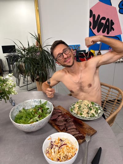 A shirtless man is posing at a table with various food items. The food includes sliced steak on a wooden board, a bowl of coleslaw, a bowl of salad, and a bowl of what appears to be cauliflower with herbs. The man is holding a fork. The setting appears to be indoors, possibly in a home. There is a potted plant nearby, along with white flowers. In the background, an abstract painting is visible. There is no information to confirm that this picture was taken in Givatayim, Israel.