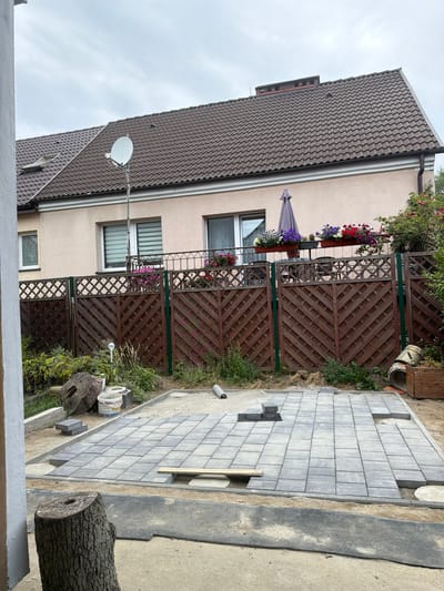 The image depicts an outdoor scene in a residential backyard, likely located in Szczecin, Poland, judging by the satellite dish commonly seen in Polish homes. In the foreground, a newly laid patio of grey interlocking pavers is under construction, with some sections incomplete. A large tree stump with a hole in its center is positioned to the left, and next to it are construction materials like buckets and a stack of pavers.
A wooden lattice fence, stained dark brown with green vertical supports, separates the yard from a neighboring property or a garden area. Behind the fence, a pale pink house with a brown tiled roof is visible. The house has windows with blinds and a satellite dish mounted on a pole in front of it. Flower boxes filled with colorful flowers adorn a balcony or railing on the house. The sky is overcast, suggesting cloudy weather. To the right, a grey umbrella is partially visible, and a wooden planter box with a plant is also present. The overall impression is one of ongoing home improvement or landscaping work.