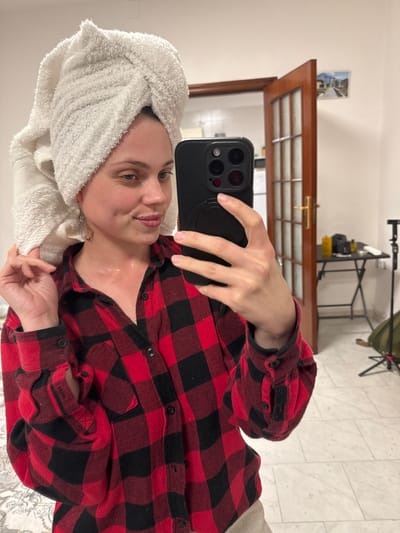 A Caucasian female subject is depicted in a mirror selfie taken indoors in Ercolano, Italy. She is positioned centrally, holding a black smartphone with her left hand to capture the image. Her hair is wrapped in a white terry cloth towel turban. She wears a red and black buffalo plaid long-sleeved button-up shirt. Her right hand is raised, lightly touching the towel on her head. Her expression shows a slight smile with lips lightly parted. Redness is visible on her neck.

The background reveals a light-colored wall and a partially open brown wooden door with multiple glass panes. A small rectangular image, possibly a postcard, depicting ancient ruins, is affixed to the wall above the door frame. Behind the open door, a black folding table is visible, holding two yellow containers, a black bag or case, and other indistinct items. To the right of the table, a black stand, possibly a tripod or light stand, is present. The floor consists of light-colored square tiles. A patterned fabric, possibly a duvet cover, is visible in the lower-left foreground.
