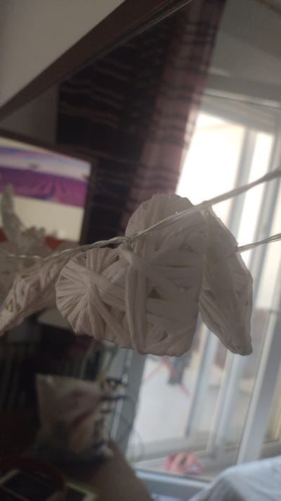 The image shows a close-up of a decorative star made of woven white material, possibly paper or straw, suspended by thin wires. The star appears to be part of a garland or string lights, with other similar star shapes visible to the left and right. The background is blurred, but suggests an indoor setting with a window showing daylight and a glimpse of a dark curtain with a striped pattern. There is also a reflection in a surface to the left, possibly a mirror or a glass table, showing a pinkish object. The scene is lit by natural light coming from the window. The image does not contain any information that would allow for its placement in Perea, Greece.