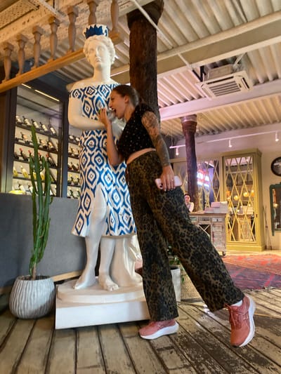 The image shows a woman in a leopard print pantsuit and a dark patterned crop top, playfully interacting with a white statue of a woman wearing a blue and white patterned dress. The statue is tall and stands on a white pedestal. The woman is leaning towards the statue, with her mouth open as if speaking to it or taking a bite, and holding a small object, possibly a piece of fruit, near its face. The scene appears to be indoors, with wooden floorboards and a ceiling with exposed beams and lighting fixtures. In the background, there are shelves stocked with wine bottles, a large cactus in a decorative pot, and some ornate furniture. The location is identified as Solnechnoye, Russia.