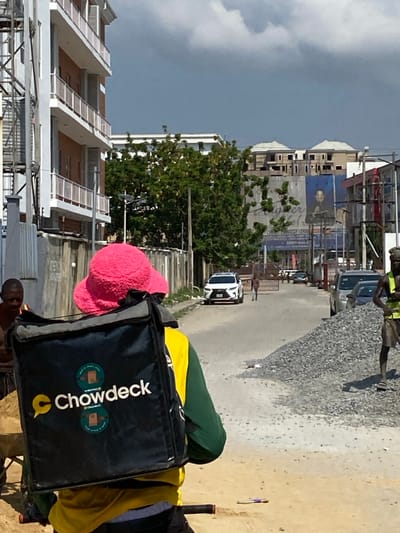 The image depicts an outdoor urban scene in Ikate, Nigeria, under a partly cloudy sky with prominent dark grey clouds. In the immediate foreground, a person is seen from behind, wearing a bright pink fluffy bucket hat, a yellow and green long-sleeved shirt, and black pants. They are carrying a large black delivery bag emblazoned with the "Chowdeck" logo in white and yellow. Bicycle handlebars are visible at the bottom of the frame, indicating the person's mode of transport. To the left of this central figure, a second individual, partially visible, stands beside a wheelbarrow filled with sand. To the right, a third person, a male in a high-visibility yellow-green vest and dark shorts, stands next to a large, conical pile of grey gravel, holding a tool (likely a shovel).
The midground features an unpaved dirt and gravel road extending into the distance. A white Lexus SUV is positioned on this road, either parked or moving slowly. Further down the road, a lone pedestrian walks away from the camera towards a distant gate or barrier. On the left, a grey concrete wall runs parallel to the road, bordering a property. Behind this wall, a multi-story building with white exterior walls, brown sections, and white-railed balconies suggests new construction or recent development. Green foliage and trees are visible beyond the wall. On the right side of the road, several dark-colored vehicles are parked, and a large billboard featuring a dark-skinned man in a suit is prominent. In the distant background, multiple unfinished buildings with light-colored roofs are visible against the horizon.