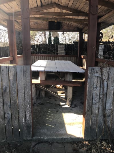 The image displays an unoccupied rustic wooden gazebo situated outdoors in Сапоцкінскі сельскі Савет, Belarus, under bright daylight. The structure features a gabled roof constructed from weathered wooden planks, supported by thick, dark brown timber posts. Decorative horseshoe motifs are carved or affixed to the top sections of at least two visible posts. A white-painted plank tabletop, showing signs of wear and cracking, is integrated into a wooden picnic table with attached benches, all constructed from substantial, irregularly shaped timber. The floor is concrete, partially covered with dry leaves and debris, and exhibits strong shadows cast by the table and the wooden structure, indicating direct sunlight from the left.
The lower half of the gazebo's perimeter is enclosed by vertical, weathered gray wooden planks, while the upper sections remain open. Inside, two hanging light fixtures are suspended from the roof; one features an ornate, possibly glass or ceramic, shade, and the other is a simpler bulb-like fixture. Beyond the gazebo, a dark wooden fence with intermittent brick pillars is visible. Behind the fence, several bare-branched deciduous trees and at least two slender, dark green coniferous trees are present. A metallic object, resembling a wheel, is visible above the fence line on the far right. Dried vines or branches cling to the exterior of the gazebo's wooden panels on the right side. No individuals or animals are present in the scene.
