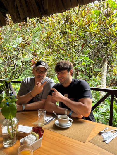 The image captures two males seated at a wooden table on an outdoor patio in Flores, Guatemala. The background is dominated by dense, lush tropical foliage, with a visible wooden railing. A portion of a thatched roof is present in the upper frame. The male on the left, an adult, wears a grey t-shirt, glasses, and a dark baseball cap. He has a silver bracelet and a light blue wristband on his left wrist. He is depicted smiling, with his right index finger raised and pointing upwards. The male on the right, a younger adult, wears a black t-shirt and a light blue wristband on his left wrist. He is also smiling, pointing his left index finger towards his right bicep. The wooden table displays, from left to right: a glass vase containing green leafy plants in water, a small glass with orange liquid, a white container holding individually wrapped condiments, an empty clear drinking glass, a white ceramic cup with a spoon resting on a saucer containing a dark beverage (likely coffee), and two sets of cutlery (fork and knife, or fork, knife, spoon) wrapped in white napkins. All dining items are placed on woven placemats.