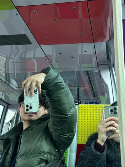 This image depicts a reflection of two individuals inside a public transit train car in Carrières-sur-Seine, France. The primary subject, positioned on the left, is a person wearing a dark green puffer jacket and glasses. They hold a silver-colored smartphone, likely an iPhone with a clear MagSafe-compatible case, horizontally to obscure their upper face, revealing only their mouth and chin. A black shoulder strap is visible across their chest. The secondary subject, partially visible on the right as another reflection, wears a dark, textured jacket over a maroon top and holds a similar silver smartphone in a clear case, also obscuring their face. The train car's interior features a gray ceiling with red paneled sections, punctuated by small embedded fixtures and a black rectangular ventilation grate. Large windows line the left side, showing a blurred exterior. On the right, a section of bright yellow, textured seating is visible, with a small portion of red seating material below it. The entire scene is captured as a reflection, likely in a large window or mirror within the train.