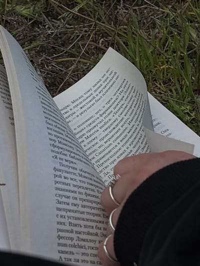 Here is a detailed description of the image:

**Scene:** An open book rests on the ground, amidst grass and other low-lying vegetation. A hand with silver rings is turning the page. The text on the open pages is in Russian. The environment appears to be outdoors.
