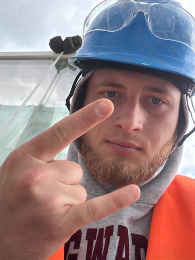 A close-up, over-the-shoulder selfie captures a young man in a blue hard hat and clear safety goggles, wearing a grey hoodie and an orange safety vest. His right hand is raised, with index and pinky fingers extended in a rock-on gesture. The man has light skin, light blue eyes, and a ginger beard and mustache. The background shows a cloudy sky and a white structure with a reflective surface, possibly part of a construction site. The image is taken outdoors, likely in Męcina, Poland, given the context of the photograph.