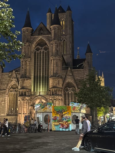 The image captures a nighttime scene in the city of Ghent, Belgium, with the imposing Saint Nicholas' Church (Sint-Niklaaskerk) as the dominant backdrop. The church, a magnificent example of Gothic architecture, is illuminated, highlighting its intricate stonework, pointed spires, and large stained-glass windows. In the foreground, several people are visible. Some are standing near a row of parked bicycles and a scooter, while others are gathered around a colorful temporary structure adorned with cartoonish depictions of animals, reminiscent of a jungle or safari theme. The ground appears to be paved, with streetlights and overhead wires suggesting an urban environment. A black car is parked on the right side of the frame, partially obscuring the view of the street. The overall atmosphere is that of a lively public space at dusk or night.