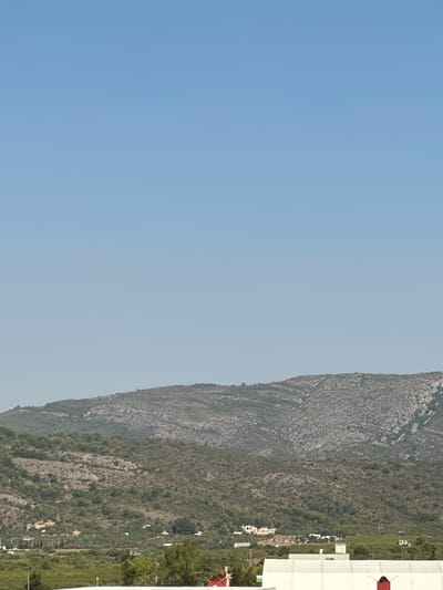 The image depicts a rural landscape with a clear blue sky. In the foreground, a portion of a large white building with a red arched opening is visible. Beyond this, a vegetated area with scattered trees and some low-lying structures can be seen. The middle ground is dominated by a hilly, sparsely wooded mountainside, characterized by rocky outcrops and dense green foliage. The background features a larger, more rugged mountain range with textured gray rock faces and sparse vegetation. The scene is located in the outskirts of Les Amplaries, Spain. There are no people visible in the image.
