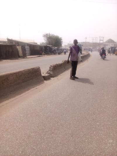 This image depicts a street scene in Tudun Murtala, Nigeria, under a bright, hazy sky indicative of dusty conditions. The foreground features a paved, light-colored asphalt road with a damaged, low concrete or mud median segmenting it. Exposed rebar is visible within the broken median.
In the mid-ground, a young male, appearing to be a teenager, stands facing slightly right and forward on the paved road, adjacent to the damaged median. He wears a faded purple and white short-sleeved shirt, dark track pants with a light stripe down the side, and sandals. A white plastic bag is slung over his left shoulder. His shadow extends to his left on the road.
Behind the individual, the street extends into the background, showing moderate traffic. Multiple motorcycles, some carrying passengers, are visible on both sides of the road. On the left side of the street, a row of low-rise commercial structures or market stalls with corrugated metal roofs and rough walls (some appearing to be mud or concrete) line the road. Several people are discernible near these structures. Further down the road on the right, more buildings are visible, including one with a distinct light-colored rounded roof. Utility poles with overhead lines are scattered across the background against the hazy sky. A green tree is visible behind the market stalls on the left.