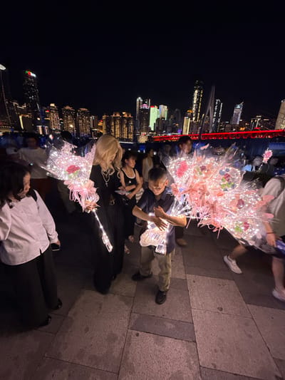 At night, a group of people are gathered outdoors on a paved walkway, with a cityscape of illuminated buildings in the background. The city skyline features tall skyscrapers of varying architectural styles and heights, some adorned with colorful lights and signage. A red illuminated bridge is visible in the mid-ground.
In the foreground, several individuals are holding large bouquets of transparent balloons filled with lights and decorative elements like flowers. A woman with blonde hair is in the center, wearing a black dress and necklaces, holding a bouquet with a single red rose. To her left, a child in a white shirt and black pants looks towards the woman. To the right of the woman, a young boy in a striped shirt and light-colored pants is holding a bouquet of balloon lights and appears to be interacting with the lights. Other people are visible in the background, some walking and others standing, some holding their phones up, possibly taking pictures or videos. The ground is paved with large, square-shaped stones. The overall atmosphere suggests a festive or celebratory event taking place in an urban environment at night.
