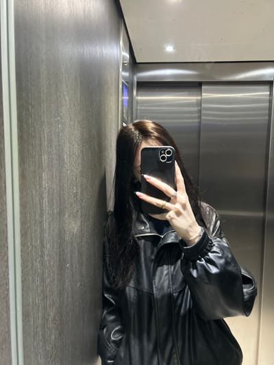 The image captures a female individual taking a mirror selfie inside an elevator. The subject's face is largely obscured by a black smartphone, held by her left hand. She wears a black leather jacket with a zippered front and ribbed cuffs, over a dark-colored top. Her long, dark hair is visible. The left hand displays long, light-colored manicured nails, a silver-toned ring on the index finger, a gold-toned ring on the middle finger, and another silver-toned ring on the ring finger. A white beaded bracelet adorns her left wrist. The smartphone is black with a multi-lens camera module accented by silver-colored embellishments. The elevator interior features a dark, horizontally grained wood or laminate panel on the left wall, and brushed stainless steel doors on the right, which reflect the subject and the opposite wall. The ceiling is white with a single visible recessed light fixture. The precise location within Cachan, France, is not visually discernible from the image.