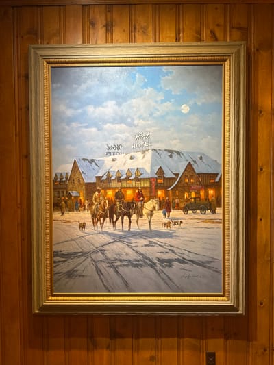 A framed painting hangs against a paneled wall. The artwork depicts a snow-covered scene at night, illuminated by a full moon. A horse-drawn carriage is arriving at a hotel; people and dogs gather around the entrance. The hotel bears the name "Word Hotel" and "Alpine". The scene evokes a sense of warmth and nostalgia. The frame is ornate, adding to the artwork's traditional and rustic aesthetic.
