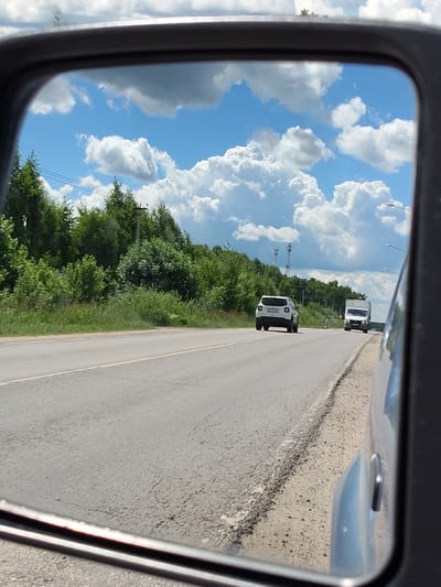 This media file shows a reflection in a car's side mirror, depicting a rural road scene in Russia, specifically near Khodaevo. The mirror reflects a clear blue sky filled with fluffy white clouds, suggesting fair weather. The road is paved with asphalt and has white lane markings. On the right side of the road, there is a gravel shoulder and the reflection of the side of the car from which the photo is taken.
Further down the road, two vehicles are visible. A white SUV is in the left lane, and a white truck is in the right lane. Both vehicles appear to be moving away from the viewer. The roadside is lined with lush green trees and bushes, indicative of a summer or late spring setting. In the background, there are power lines and utility poles, and two cell towers can be seen in the distance. Streetlights are also visible along the side of the road. The overall scene conveys a sense of travel on a pleasant day. There is no visible text in the image.
