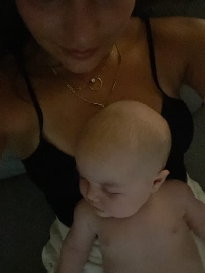 The image depicts a close-up, overhead view of a baby being held by an adult. The adult is wearing a black top and a gold necklace with a small pearl pendant. The baby, who appears to be very young, is napping with its eyes closed and is partially undressed. The setting is indoors, but no specific details of the environment are discernible due to the close framing and lighting. The image does not provide any information about the location in Tonbridge and Malling, United Kingdom.