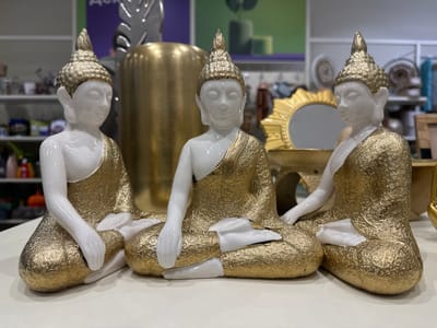 The image shows three seated Buddha statues, presumably decorative items for sale, on a table or shelf. They are predominantly white with gold detailing on their robes and headdresses. The statues are in a meditative pose, with crossed legs and hands resting in their laps.

Behind the statues are other decorative items, including a gold-colored cylindrical vase, a metallic leaf-shaped ornament, and a gold sunburst mirror. The background is blurred, suggesting the setting is a shop or retail environment. There is shelving visible with various items on display. There is no information in the image to locate this store in the city of Kudrovo, Russia.