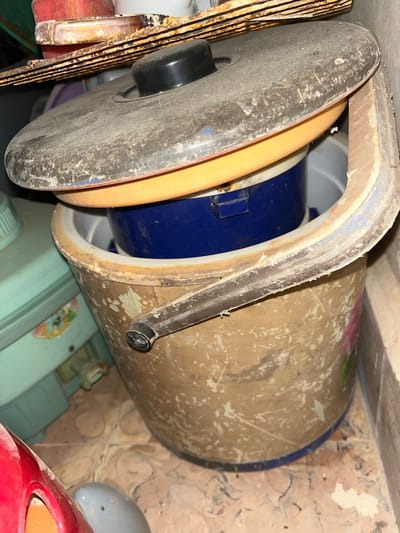 The image shows a close-up of a dusty, old, brown bucket with a blue container and a yellow lid inside, topped by a larger, dark, dusty lid with a black knob. The bucket has a metal handle with a black grip. In the background, a teal-colored container with floral designs is visible, as is a portion of a red object and a gray object in the foreground. The scene appears to be an interior space, possibly a storage area, with a tiled floor and a brick wall. The overall condition of the objects suggests they are old and have been exposed to dust and dirt. The location is described as being in Kota Kuala Simpang, Indonesia.