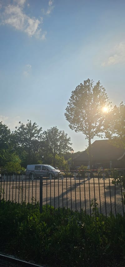 The image depicts a sun-drenched scene on a paved road, likely in Groenlo, Netherlands, with a black metal fence in the foreground. A silver van is driving on the road, casting a shadow. Beyond the fence, lush green foliage and trees are visible, with a large tree to the right of the frame partially obscuring the bright sun, which creates lens flare. In the background, a building with a dark roof and brickwork is visible, along with a lamppost and other trees. The sky is blue with scattered clouds.