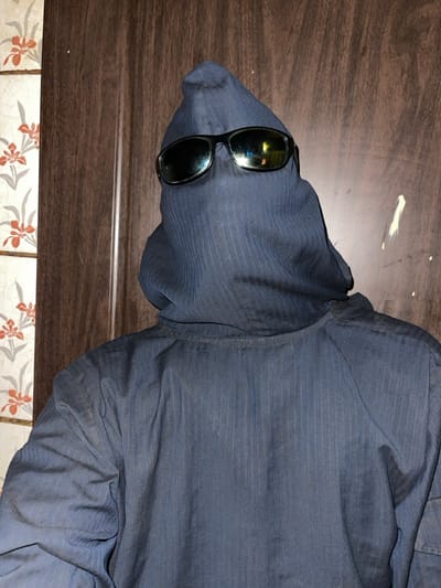 A dark blue button-down shirt lies on a surface transitioning from brown wood to tiled floral patterns. The shirt's left sleeve is bundled, concealing a head-like shape. Dark sunglasses sit on this form, mimicking eyes. A small dab of white is on the wood surface above the "head". The shirt's fabric has subtle, fine lines, adding texture to its appearance.