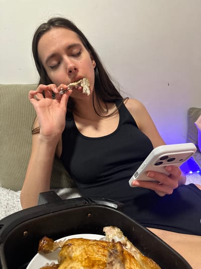 An indoor scene in Corby, United Kingdom, depicts a female individual eating cooked poultry. The subject, possessing long dark hair, wears a black ribbed tank top and is seated on a light-colored sofa or cushion. Her eyes are closed or cast downwards as she brings a bone, stripped of most meat but with some residue, to her mouth with her right hand. Her left hand holds a white smartphone. In the foreground, a black air fryer contains a white plate holding additional pieces of cooked, golden-brown poultry, some of which appear partially consumed. The background features a plain, light-colored wall, with blue string lights visible to the right.