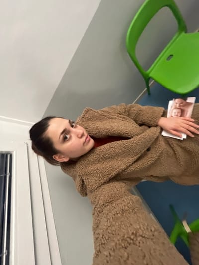 A woman with dark hair pulled back and wearing a brown, plush coat sits in a room. She holds a printed photo, possibly of a baby, in her left hand. A bright green chair is partially visible beside her. The room has white walls and a window with blinds at the bottom of the frame. The image appears to be rotated, causing the perspective to be upside down. The woman looks toward the camera with a slight upward gaze.
