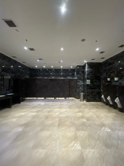 The image depicts an interior view, possibly a modern hallway or lobby, with a striking contrast between dark, glossy marble-tiled walls and a bright, textured wall. The scene is oriented vertically, with the viewpoint looking upwards towards a ceiling that features several white light fixtures and what appear to be ventilation grilles.
On the left side of the image, a white wall is punctuated by square vents and circular light fixtures, suggesting a functional but clean design. The wall is smooth and reflects light.
The central and right sections are dominated by dark, highly polished marble with prominent veining, creating a dramatic and luxurious aesthetic. This dark marble extends to the floor and appears to form part of a staircase or elevated platform. Several white, elongated light fixtures are mounted on the ceiling above this dark section.
To the far right, a wall is covered in large, light-colored tiles that have a textured, almost translucent quality, with visible patterns that create a play of light and shadow. This creates a softer, more ethereal atmosphere compared to the dark marble.
There is no visible activity or people in the frame, suggesting the image was taken in an empty space. The lighting is bright, indicating it's likely daytime or the space is well-lit internally. The overall impression is one of modern architectural design, emphasizing luxurious materials and contrasting textures. The location context of Ahmedabad, India, is noted but not directly evident from visual cues within the image itself. There is no visible text in the image.