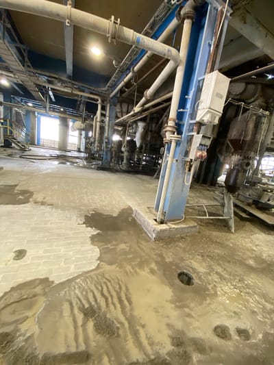 This image depicts the interior of an industrial facility, likely a manufacturing plant or processing center, with a focus on machinery and infrastructure. The scene is characterized by a large amount of piping, large industrial equipment with gears and housings, and a complex network of walkways and platforms. The floor appears to be a light-colored brick, covered in areas by a wet, muddy substance, suggesting a recent event involving water or other liquids. There are several drains visible in the floor, some covered and some open.

The lighting in the image is somewhat dim, with a bright light source originating from a window or opening in the background, suggesting it might be daytime. The overall impression is one of a functional, perhaps older, industrial environment. There is no visible human activity or indication of people in the frame. The materials are predominantly metal and brick, with a significant presence of water or sludge on the floor. The image appears to be taken with a wide-angle lens, giving a sweeping view of the space. The crops highlight the details of the machinery, the construction of the piping systems, and the texture of the floor surface with its wet and muddy patches. There is no visible text in the image.