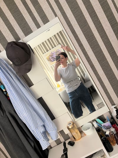 A woman is taking a mirror selfie in a room. She is wearing a striped t-shirt and blue jeans. Her hair is dark and pulled back. She is making a peace sign with her left hand. In the reflection, a framed picture of lavender can be seen on the wall. Next to the mirror, there is a rack with clothes hanging on it, including a striped blue and white shirt and a dark gray garment. On a shelf below, there are various items such as bottles of perfume, a golden cylindrical object, a white dish with paper clips, and a black smartwatch. The room has striped wallpaper in shades of gray and beige. There is no indication that this scene is located in Sochi, Russia.