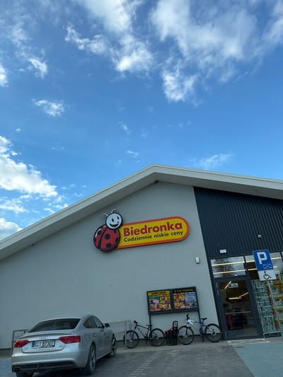 The image shows the exterior of a Biedronka store in Męcina, Poland. The store has a modern facade with a slanted roof and dark vertical siding. A large Biedronka logo, featuring a ladybug, is prominently displayed on the wall. In front of the store, a silver Audi sedan is parked. Adjacent to the car, there are several bicycles. A glass entrance door leads into the store, which appears to be well-stocked with products, including toilet paper visible through the glass. The sky is blue with scattered white clouds.