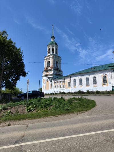 This is a photograph of a church in the village of Mostovoe, Russia, under a clear blue sky with scattered clouds. The church is white with yellow trim, featuring a tall bell tower topped with a green spire and a cross. The main building has a green roof and a series of arched windows. In the foreground, a black sedan is parked on the side of a paved road. A traffic sign with a circle and a number inside is visible near the car. The surrounding area is covered in green grass and vegetation, with a dirt embankment sloping down to the road. Trees are present on the left side of the image. Several birds are visible flying in the sky.