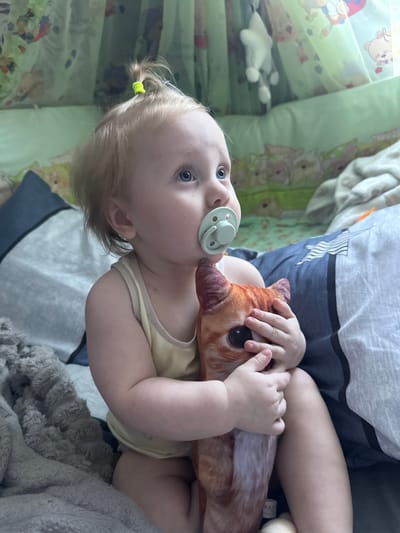 In Sayanogorsk, Russia, a Caucasian infant with blonde hair, styled in a small ponytail, sits on a bed. The infant wears a yellow tank top and has a light blue pacifier in its mouth. The infant clutches a plush toy shaped like a cat's head. The infant's eyes are open, and it looks upwards and to the right. The bed is covered with a blue and grey patterned duvet, and a grey fuzzy blanket is visible on the left. In the background, a green crib liner with a pattern of cartoon animals, including bears, is visible.