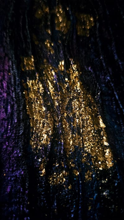 The image depicts a close-up, abstract view of a textured surface with shimmering gold and purple elements against a dark background. The texture appears to be fabric or a similar material with a velvety or crinkled quality. The gold and purple flecks catch the light, creating a sparkling or glittery effect. The lighting is dramatic, with strong contrasts between the illuminated areas and the deep shadows, suggesting artificial lighting or a specific light source. The overall impression is one of luxury, mystery, or a festive atmosphere. There are no discernible people, animals, or recognizable objects in the scene. The location in the city of Kostenets, Bulgaria, is not visually represented in the image; this is an abstract detail without any visual cues to support it.
