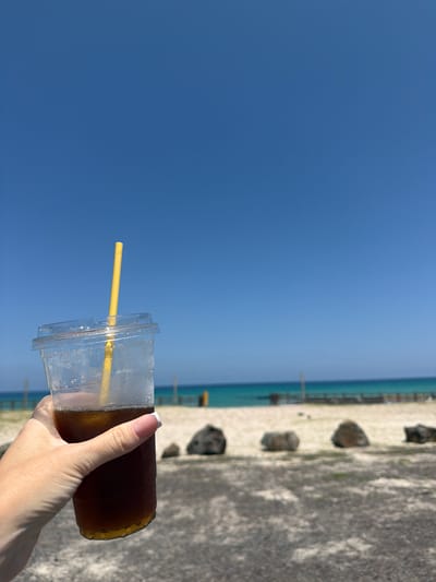 A person's hand, with a French manicure, holds a clear plastic cup filled with a dark beverage, likely iced coffee, and a yellow straw. The cup is partially filled, with condensation visible on the exterior. The background shows a bright blue sky dominating the left side of the frame, transitioning to a blurred view of a beach with sand, rocks, and the ocean on the right. The ocean appears calm with a thin, distant horizon line. The image is oriented vertically, suggesting the photo was taken with the phone held in portrait mode. The lighting indicates it is daytime, likely sunny, creating a relaxed and possibly vacation-like atmosphere. The location is specified as Jeju, South Korea, hinting at a coastal or beachside setting. There is no visible text in the image.