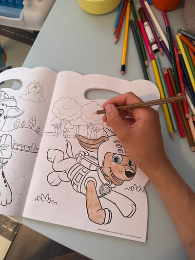A hand is coloring in a Paw Patrol coloring book. The coloring book is open to two pages, with drawings of characters from the show. The hand is holding a light brown colored pencil and is coloring in the fur of one of the characters. There are other colored pencils scattered on the table next to the coloring book. The scene appears to be a domestic setting, likely a home. The image does not contain any information that would allow for the determination of its location within Gatineau, Canada.