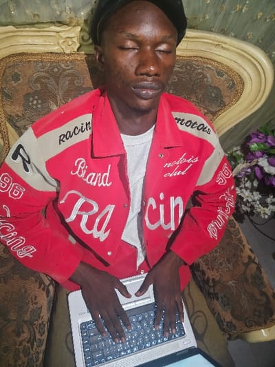 A man with dark skin lies on a patterned couch with a laptop resting on his lap. He wears a red jacket with "Racing" printed on the sleeves. His eyes are closed, suggesting he might be sleeping. His hands rest on the laptop's keyboard, indicating he may have fallen asleep while using the computer. Flowers are visible in the background. The couch appears ornate with a decorative pattern.