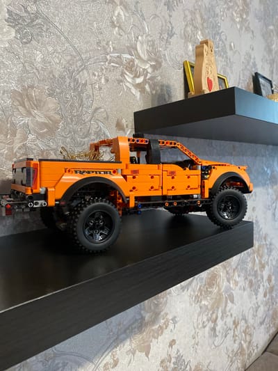 An orange LEGO Technic Ford Raptor pickup truck is displayed on a black shelf against a wall with floral wallpaper. The truck is detailed, featuring visible gears, suspension components, and the "RAPTOR" logo on the side. In the background, on a higher shelf, are a few decorative items, including a wooden figurine with a heart, a framed picture, and another framed item. The scene appears to be indoors, possibly in a residential setting, and there is no indication of the specific location within the city of Berëzovaya Roscha, Russia.