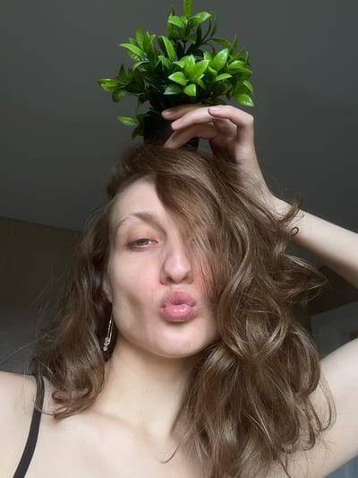 The image presents a close-up, rotated portrait of an individual, likely female, in an indoor setting in Podshivalovo, Russia. The person is depicted as if lying horizontally, with their head positioned towards the right of the frame and their body extending to the left.
The subject has fair skin and voluminous, wavy light brown hair, styled with subtle lighter highlights. Their face is partially visible, dominated by a playful expression featuring puckered, glossy pink lips in a kissing pose. The left eye, a striking green or hazel, is open and looks directly at the camera, while the nose is also visible. Some strands of hair gently fall across the forehead.
A slender left hand rests casually atop the person's head, cradling a small, artificial plant with numerous bright green leaves in a dark container. Another arm is partially seen in the upper left, also appearing to be resting on the head amidst the hair. On the right side of the frame, near the neck, a delicate earring or hair accessory adorned with three small, sparkling clear gems is visible. The person is wearing a dark, possibly black, clothing strap that hints at a top or undergarment.
The background is a uniform, plain dark gray wall, indicating an uncluttered indoor environment. The lighting is soft and diffused, creating a gentle play of light and shadow that enhances the subject's features. There is no visible text within the photograph. The overall mood is intimate and casual, capturing a moment of relaxed posing.