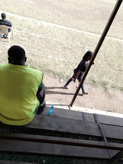 The image portrays an outdoor sports facility in Nakuru, Kenya, under bright sunlight. Three individuals are visible. In the immediate foreground, viewed from behind, a dark-skinned adult male wearing a bright yellow sleeveless vest and dark shorts sits on a weathered wooden bench. A blue plastic water bottle with a white cap rests on the bench to his right. A dark, angled metal support beam partially obstructs the view toward the right mid-ground. To the left of this beam, a dark-skinned individual wearing a black cap, black t-shirt, dark blue jeans, and light-colored sneakers walks leftward on a dirt perimeter track. This individual holds a small, reddish-orange rectangular object in their right hand. Further into the background on the left, a dark-skinned adult male wearing a dark cap and a black t-shirt with a white "Aquamist" logo on the back is seated on a white plastic monobloc chair directly on the field. The ground is a dry, sparsely vegetated field with light-brown grass and visible patches of bare earth. White painted lines are discernible on the field, indicating sports markings. Strong shadows are cast by the individuals and objects, consistent with direct daylight.
