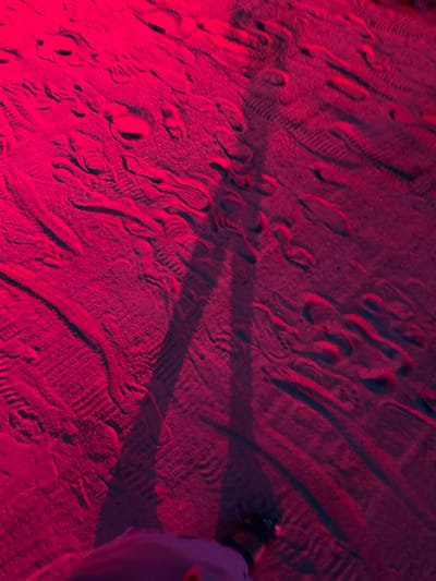 The image shows a close-up, low-angle view of a sandy surface illuminated by a bright pink light. The sand is textured with numerous footprints, some appearing as distinct imprints and others as blurred trails. There is a long, dark shadow cast across the sand, suggesting a person standing in the light source's position. The shadow appears to be that of a human figure, with discernible leg and foot shapes. In the lower right corner of the frame, a dark object, possibly a shoe or bag, is visible at the edge of the sand. The overall scene suggests an outdoor environment, potentially a beach or sandy pathway in Ko Pha-ngan District, Thailand, during nighttime or under artificial lighting.