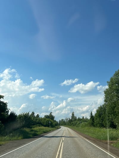 The image depicts a paved road in a rural area, likely in or near the village of Garevaya, Russia. The road is divided by double yellow lines and curves slightly to the left, disappearing into the distance. Lush green vegetation, including trees and tall grass, lines both sides of the road. The sky above is a vibrant blue, dotted with scattered white cumulus clouds. The overall impression is one of a clear, sunny day in a natural, unpopulated setting. There are no people or vehicles visible in the frame.