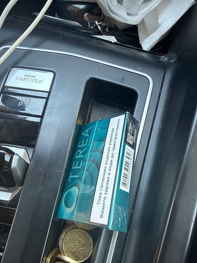 A pack of Terea Turquoise tobacco sticks for IQOS is resting in the center console storage compartment of a vehicle. The console also contains several euro coins. The "ENGINE START/STOP" button is visible above the storage compartment. The environment appears to be the interior of a car parked outdoors, possibly in a sunny location. The image is taken from a slightly elevated angle, looking down into the console. No people are visible.