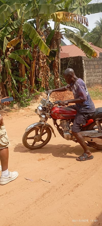 The scene depicts an outdoor interaction on an unpaved, reddish-brown dirt road in Abagana, Nigeria, recorded on 03/12/2025 at 11:01. In the center-right, a dark-skinned adult male with a bald head is seated on a red motorcycle, oriented towards the left. He wears a blue and white patterned short-sleeved shirt, dark shorts, and dark sandals. His right foot is on the ground, and both hands are on the motorcycle's chrome handlebars. The motorcycle features a chrome front fender, multiple chrome headlights, and a five-spoke front wheel. Markings resembling "OS" or "GS" are visible on the motorcycle's fuel tank. To the far left, the lower body of a second dark-skinned adult is partially visible, wearing khaki shorts, white socks, and white shoes. This individual's left hand holds a black smartphone horizontally. The background is characterized by dense tropical vegetation, primarily large banana or plantain trees with green and dried brown leaves. Behind the vegetation and to the right, a building with a reddish-brown corrugated roof and a light-colored brick wall are visible. The dirt ground exhibits tire tracks and footprints.