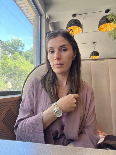 A woman with long, dark brown hair is seated indoors, looking directly at the viewer with a neutral expression. She is wearing a dusty rose or light purple long-sleeved top made of a textured, crinkled fabric. A silver and gold-toned wristwatch with a white dial and chronographs is visible on her left wrist. Her left hand is resting, while her right hand is positioned near her shoulder. Glasses are perched on top of her head.

The setting appears to be a cafe or restaurant, featuring plush, light brown upholstered booth seating. The wall behind her is decorated with several black, geometric bowl-shaped fixtures that have golden interiors; some of these contain green foliage, resembling modern wall sconces or planters. A large window to her left reveals a bright, clear blue sky and lush green trees outside, indicating a sunny daytime. A small white security camera is mounted above the window. A dark wooden surface with a geometric pattern is visible in the foreground, likely part of a table or chair. No visible text is present in the image.