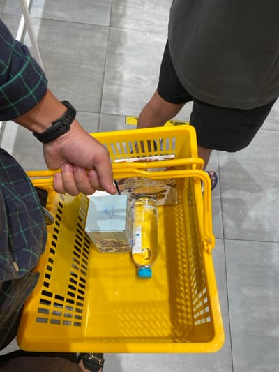 An overhead shot captures a person holding a bright yellow shopping basket inside what appears to be a store or supermarket in Bekasi Regency, Indonesia. The hand holding the basket belongs to an individual wearing a dark blue and green plaid shirt and a black digital watch on their left wrist. Partially visible next to them are the legs and lower torso of another person, clad in dark shorts and blue flip-flops.

Inside the yellow plastic basket, which has a slotted design, are a few items. A clear plastic bottle with a light blue cap, possibly containing water or cooking oil, rests on its side. Below it is a small, light blue box with a white label featuring an ornate design, possibly floral, and some unreadable text. Another flat item, resembling a magazine or packaged food, is also visible. Text that appears to read "Semprong" is somewhat discernible on this item. The floor is tiled with light grey squares. The overall lighting suggests an indoor, daytime setting.