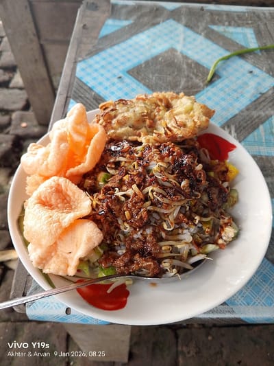 A plate of Indonesian Gado-Gado sits on a weathered table with a blue and white checkered pattern. The dish features rice, bean sprouts, cucumbers, and fried tofu, lavishly drizzled with a dark, savory peanut sauce. Two orange shrimp crackers lean against the salad. A spoon rests on the plate's edge.