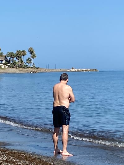 An adult male, viewed from the rear, stands at the edge of the Mediterranean Sea on a beach in Estepona, Spain. The man is wearing black swim shorts and his upper body is bare, revealing skin. He has his arms crossed in front of him. The water is calm and blue, with small waves lapping at the shore. The beach is composed of small pebbles and sand. In the background, palm trees and a building are visible on the land, with a breakwater extending into the sea. The sky is clear and blue.