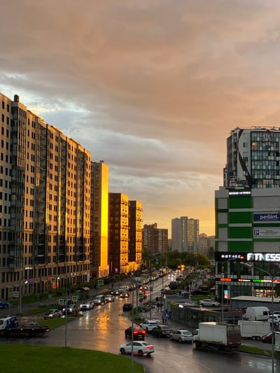 This media file captures a street view in Kudrovo, Russia, during what appears to be sunset. The sky is filled with dramatic, cloudy formations, tinged with the warm hues of the setting sun, creating a striking contrast with the urban environment.

Dominating the scene are several modern, multi-story buildings. One building in the foreground features a textured, patterned facade and reflective windows that catch the golden light of sunset. Adjacent to it, another building is clad in green and white panels, with prominent signage. Visible text on this building includes "pedant.", "АРЕНДА ПАРКОВОЧНЫХ МЕСТ +7 981 715-42-12", and "ФИТНЕС FITNESS". Another sign reads "ВКУСНО-И ТОЧКА", and "оптика".

Below the buildings, a busy street is visible, lined with a steady stream of cars, suggesting rush hour or a typical evening traffic flow. The wet road surface indicates recent rain, with reflections of headlights and streetlights shimmering on the asphalt. A few pedestrians are present, navigating the sidewalks, and crossing the street at designated crosswalks marked with blue and yellow signs. The presence of trucks and various passenger vehicles suggests commercial and residential activity in the area. The overall impression is one of a bustling, modern city district at the end of the day, bathed in the atmospheric light of sunset and a recent rain shower.