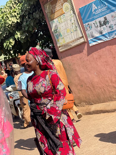 The image depicts a bustling outdoor scene in Samaru Ultra Modern Market, Samaru, Zaria, Kaduna State, Nigeria. In the foreground, a woman with dark skin wears a vibrant red dress with a yellow, white, and black leaf-like pattern, a matching headtie, and large hoop earrings. She carries a brown leather shoulder bag with a gold 'B' emblem and has a black fabric wrapped around her waist. She is smiling and looking to her right.
Behind her, several individuals are visible: a man in a light brown traditional garment and a dark cap, another man in a light blue t-shirt and a light brown woven cap, and other indistinct figures suggesting a crowd. A white and silver motorcycle is partially visible in the midground to the left.
The background on the right consists of a salmon-pink textured wall with two signs mounted. The left sign, framed in wood, advertises "Arsalan STORES" for "High Quality Skincare & Haircare Products." The right sign is a blue banner for "AL-MUBAARAK VARIETY MALL," offering "AFFORDABLE PHONE AND ACCESSORIES, HOUSEHOLD EQUIPMENT AND KITCHEN ITEMS," and explicitly states its location as "Samaru Ultra Modern Market... Samaru, Zaria, Kaduna State." Dense green tree foliage is visible in the upper left background. The scene is brightly illuminated by direct sunlight, casting shadows on the wall and ground, indicating daytime activity in a commercial area.